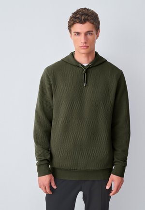 Junger Mann mit kurzen lockigen Haaren, der einen strukturierten dunkelgrünen Hoodie und dunkle Hosen trägt, steht vor einem schlichten hellen Hintergrund.