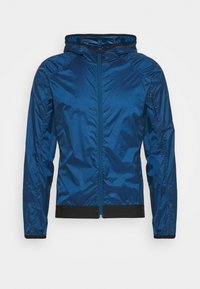 Lätt blå vindjacka för män med huva, framdragkedja, elastiska muddar och svarta sömmar mot en vit bakgrund.