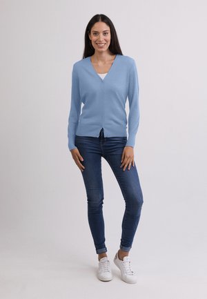Hellblaues Cardigan mit V-Ausschnitt, Knopfleiste vorne und langen Ärmeln, kombiniert mit enganliegenden dunklen Jeans und weißen Turnschuhen. Glatte Textur.