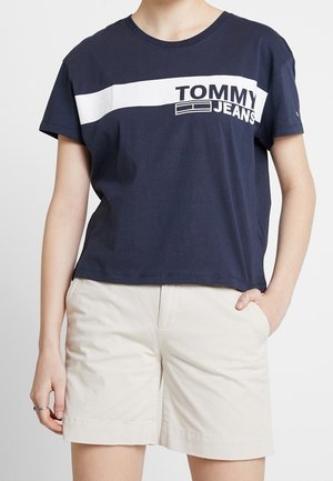 Kobieta w granatowym t-shircie Tommy Jeans z białym paskiem i beżowych szortach, pozująca z jedną ręką w kieszeni.