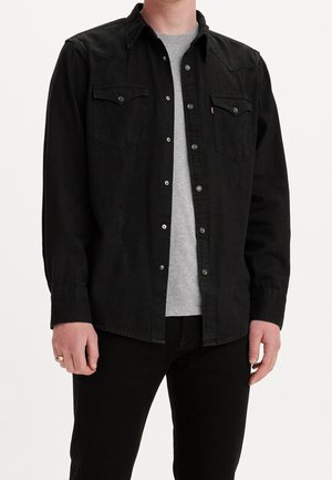 Chemise en denim noir avec des boutons à pression, deux poches poitrine, un empiècement pointu et des manches longues, superposée sur un t-shirt gris.
