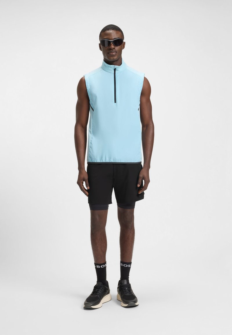 Muž v bezrukávovém světle modrém topu na zip, černých šortkách, černých ponožkách, černých teniskách a tmavých slunečních brýlích stojí před jednobarevným pozadím.