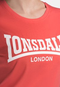 Red cotton T-shirt featuring a white curved text "Lonsdale London" prominently displayed across the chest, with a smooth texture.