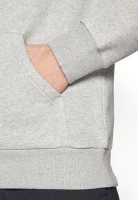 Haut à capuche gris avec une poche kangourou et des poignets côtelés. Matériau doux avec une texture brossée, mettant en valeur un détail de coutures le long des seams.