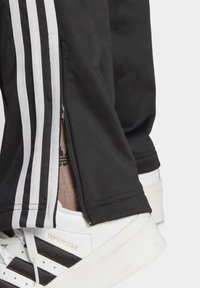 Svarta idrottsbyxor med tre vita ränder på sidorna och en dragkedja vid fållen, kombinerat med vita sneakers med svarta detaljer.