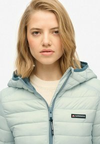 Lyseblå pufferjakke med hætte, quiltet tekstur, lynlås foran og et logo-patch på venstre side. Blød beige sweater under.