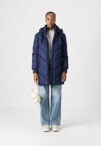 Doudoune marine avec surpiqûres en chevron, capuche et bretelles noires ; associée à un jean large bleu clair et des baskets blanches, tenant un petit sac.