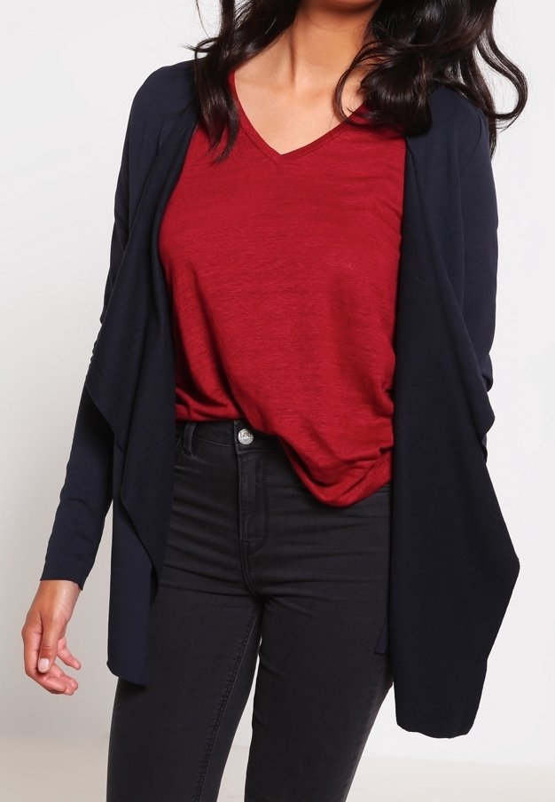 Femme portant un t-shirt rouge à col en V, un jean noir taille haute et un cardigan bleu marine ample ouvert devant sur un fond uni.
