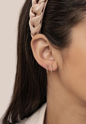 Goldene Creolen mit kleinen grünen Edelsteinen, präsentiert neben einem geflochtenen beigen Haarband, mit langen dunklen Haaren im Hintergrund.