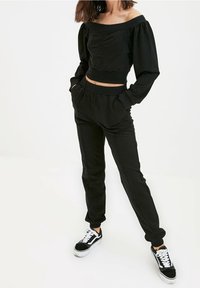 Schwarzes, kürzeres Top ohne Träger mit puffigen Ärmeln und vorderer Tasche, kombiniert mit schwarzen, elastischen Hosen. Getragen mit schwarz-weißen Sneakers.