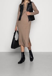 Robe tricot brun ajustée avec des fentes latérales, superposée sous un gilet en cuir noir. Associée à des bottines à talons noires et un sac à bandoulière noir.