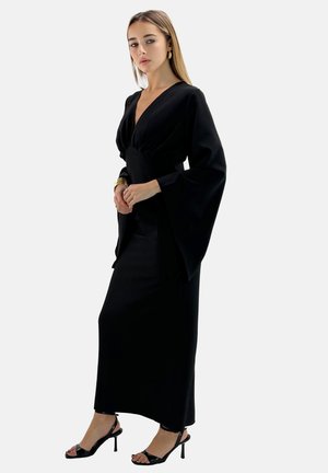 Robe noire longue avec un décolleté en V profond et des manches évasées, fabriquée en tissu lisse. Associée à des talons noirs à lanières. Design simple et épuré.