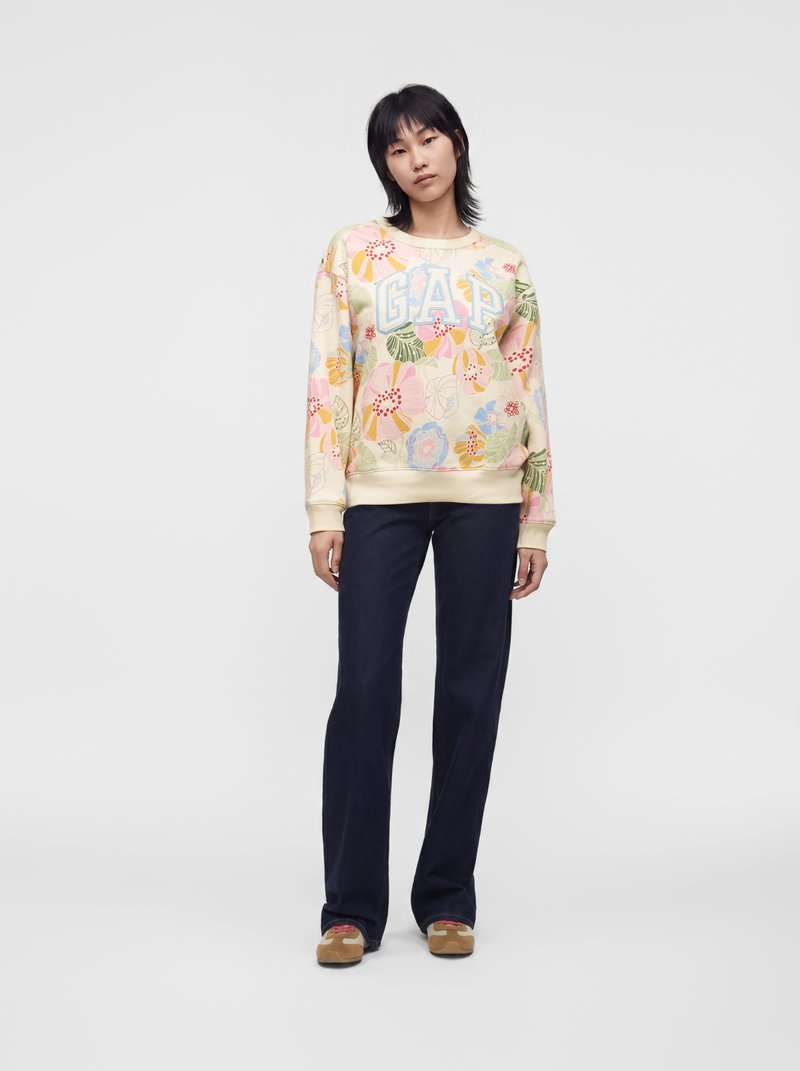 Personne debout face à l'objectif, portant un sweat GAP à fleurs, un pantalon sombre et des chaussures beiges sur un fond clair et uni.