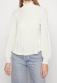 Weißes Langarmoberteil mit strukturiertem, gefälteltem Design, hohem, gerafftem Kragen und elastischen Bündchen, kombiniert mit hellblauer Jeans.