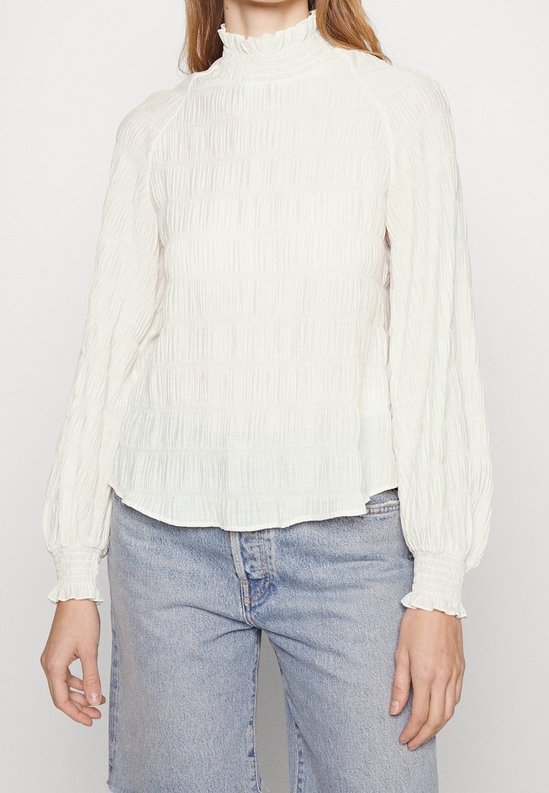 Weißes Langarmoberteil mit strukturiertem, gefälteltem Design, hohem, gerafftem Kragen und elastischen Bündchen, kombiniert mit hellblauer Jeans.