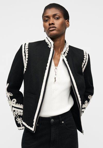 Γυναίκα που φοράει μαύρο μπουφάν με λευκή κεντημένη διακόσμηση πάνω από λευκό μπλουζάκι και μαύρο παντελόνι, στέκεται μπροστά σε ένα απλό φωτεινό φόντο.