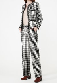 Costume motif pied-de-poule noir et blanc avec une veste courte, un pantalon long et des franges texturées. Porté avec des chaussures à talons bordeaux.