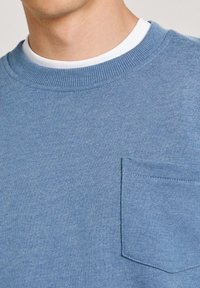 Mėlynas apvalios iškirptės džemperis, pagamintas iš tekstūruotos medžiagos, su krūtinės kišene ir raukšlėtu apykaklės kraštu. Paprastas ir minimalistinis dizainas.