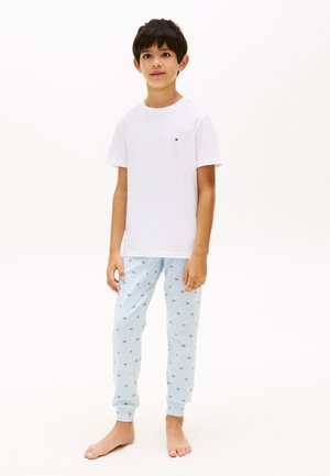 Gutt som står barfot iført en hvit kortermet skjorte og lys blå mønstrede pysjamasbukser mot en ensfarget hvit bakgrunn.