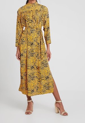 Abito leopardato giallo e nero, con maniche lunghe, scollatura arricciata, vita sfiancata e spacco laterale. Abbinato a tacchi a blocco beige.
