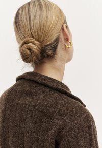 Brauner Wollcardigan mit strukturiertem Finish und umgeschlagenem Kragen. Das Modell trägt einen goldenen, abstrakt geformten Creolenohrring. Die Haare sind zu einem Dutt frisiert.
