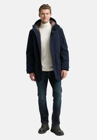 Marine parka-jack met verstelbare capuchon, met bont gevoerde kraag, knoop- en ritssluiting, gecombineerd met blauwe jeans en zwarte laarzen.