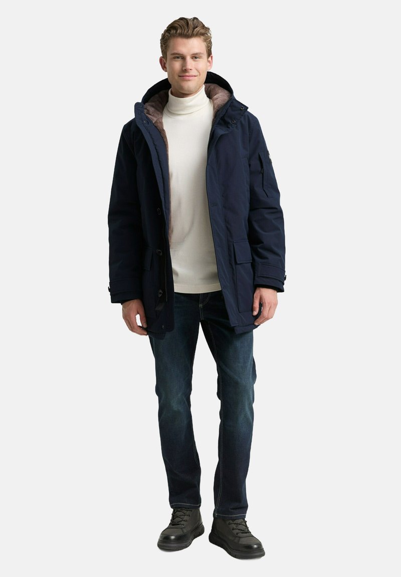 Marine parka-jack met verstelbare capuchon, met bont gevoerde kraag, knoop- en ritssluiting, gecombineerd met blauwe jeans en zwarte laarzen.