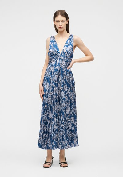 Femme debout, main sur la hanche, portant une robe maxi bleue sans manches avec un motif floral blanc et des talons noirs à lanières, sur fond uni.