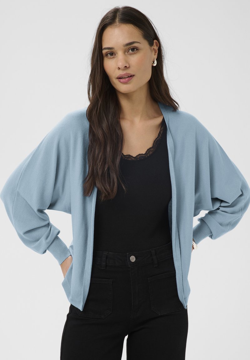 Pikkade tumejate juustega naine, kellel on seljas hele sinine avatud kardigan, must pitsäärisega pluus ja mustad kõrge vöökohaga püksid, seisab käed taskutes.