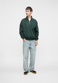 Tmavě zelená mikina na zip s uvolněným střihem, nošená přes bílý tričko. Světle modré džíny a černé tenisky s gumovou podrážkou.