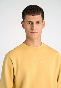 Felpa gialla realizzata in materiale morbido con scollatura a girocollo. Presenta il logo ricamato "LINDBERGH" sul davanti. Tessuto liscio.