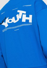 Niebieska bluza z dżerseju, z dużym białym napisem "lost YOUTH" na plecach oraz dodatkowymi mniejszymi tekstami w różnych czcionkach.