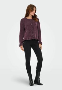 Cardigan texturé violet foncé avec des boutons à l'avant et un ourlet froncé, associé à un jean noir ajusté et des bottines, se tenant devant un fond uni.