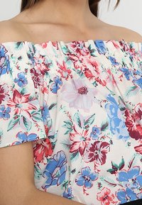 Donna con una blusa bianca senza spalle, decorata con un motivo floreale rosso, blu e viola, e un collo arricciato.