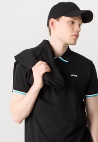 Czarna koszulka polo z krótkimi rękawami, akcentami kołnierza w paski w kolorze aqua i białym oraz logo na piersi. Model trzyma czarną bluzę.