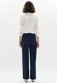 Femme aux cheveux bouclés à mi-longueur portant un chemisier blanc et un pantalon large bleu marine, se tenant de dos sur un fond blanc.
