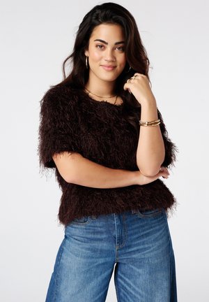 Jonge vrouw met lang donker haar, draagt een pluizige donkerbruine top en blauwe spijkerbroek, glimlacht met haar hand rustend bij haar gezicht.