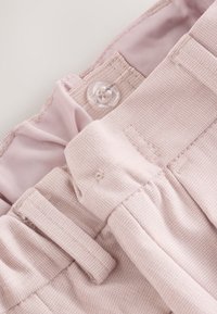 Gros plan sur la ceinture d'un pantalon rose clair montrant un bouton transparent, une boucle de ceinture et un détail de tissu froncé.