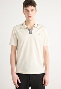 Kurzarm-Polo-Shirt aus off-weißem Baumwollstoff. Mit einem schwarz-weißen gestreiften Kragen und einem kleinen Logo auf der Brust. Einfaches und sauberes Design.
