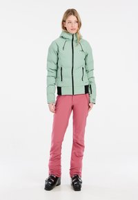Lichtgroene pufferjack met een hoge kraag, voorrits en zwarte accenten, gecombineerd met roze skibroek en zwarte skischoenen.