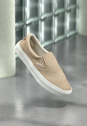 Beiger Slip-On-Sneaker mit weißer Sohle und Hilfiger-Logo, schwebt über einem grauen Boden vor einer Wand aus Glasbausteinen.