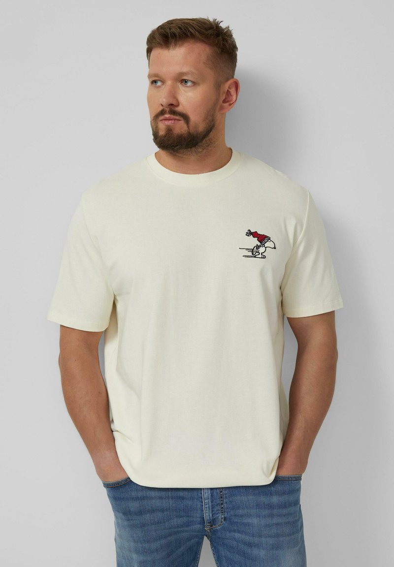 Man with beard wearing a cream t-shirt with a small cartoon figure in red above left chest, standing with hands in pockets against gray background.