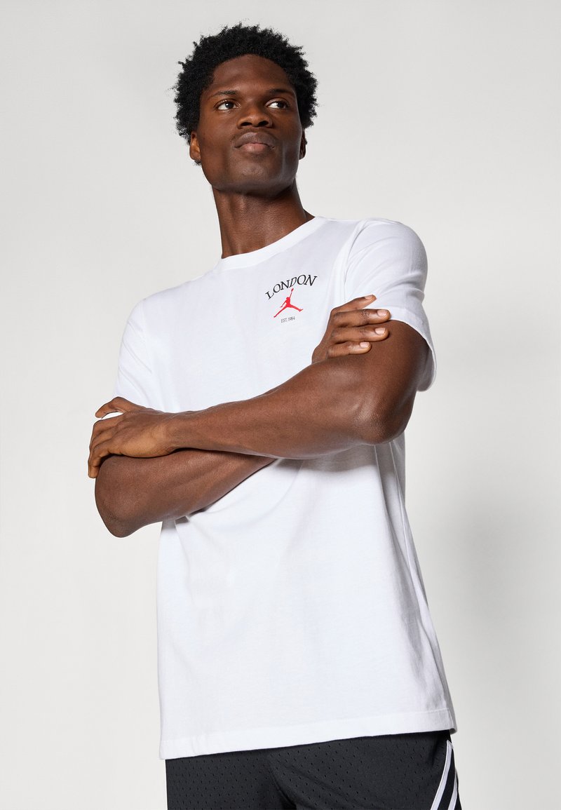 Homme portant un t-shirt blanc avec "London" et un logo rouge, les bras croisés, regardant légèrement vers le haut, vêtu de shorts noirs avec des rayures.