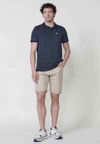 Námornícky polo tričko s textúrovaným vzorom, béžové krátke nohavice s vyhrnutými okrajmi a šedé športové topánky s námorníckymi a bielymi akcentmi. Model stojí neformálne.