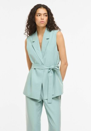 Donna con capelli ricci che indossa un blazer azzurro chiaro senza maniche con cintura e pantaloni coordinati, guarda di lato con le mani dietro la schiena.