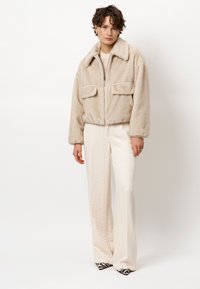 Beige nepbont jas met een ritssluiting aan de voorkant, een oversized kraag en twee voorkeuzes, gecombineerd met crèmekleurige broeken en gebloemde hakken.