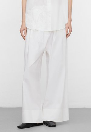 Persoon draagt losse witte wijde broek, bijpassend wit shirt en zwarte puntige schoenen, staand op een grijze vloer.