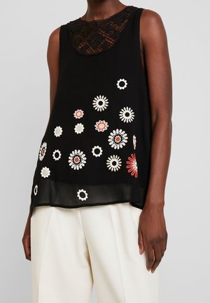 Haut noir sans manches avec un col en dentelle et des motifs floraux blancs et rouges, porté avec un pantalon blanc ample.