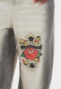 Jeans de mezclilla gris con un gran parche bordado colorido de una rosa y una pancarta que dice "EN MEMORIA DE CUANDO ME IMPORTABA". Textura desgastada visible.