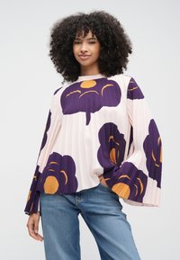 Halvány rózsaszín blúz lila és narancssárga virágmintákkal, pliszírozott kialakítással és széles harang ujjaival. Világoskék farmerrel párosítva.